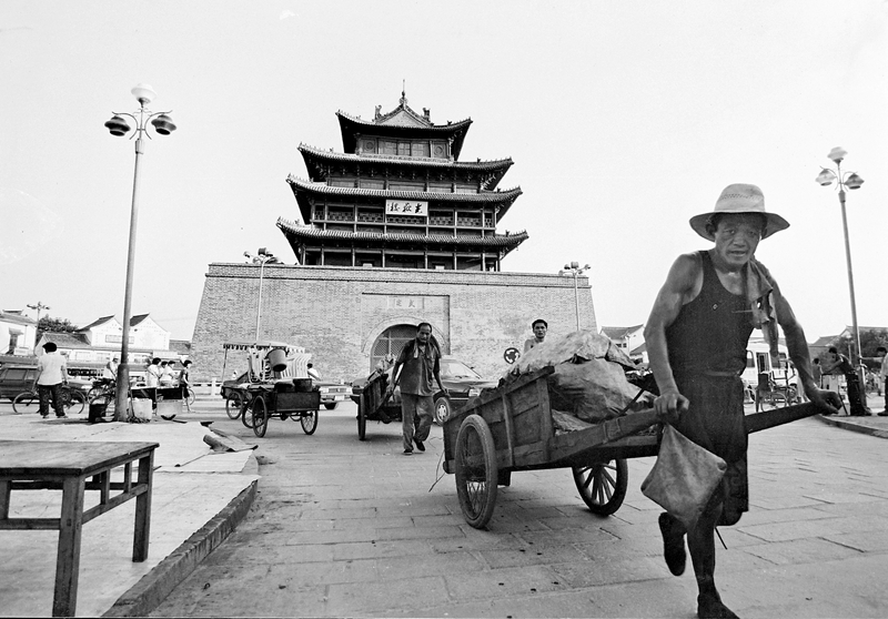 19、《古樓下的板車(chē)》1996年8月,聊城市區(qū)光岳樓前,兩位男子拉著板車(chē)依次前行。在他們身后,小轎車(chē)慢慢行駛著。在上世紀(jì)90年代,進(jìn)城的商販運(yùn)送貨物大多采用這樣的板車(chē)。(林虎).JPG